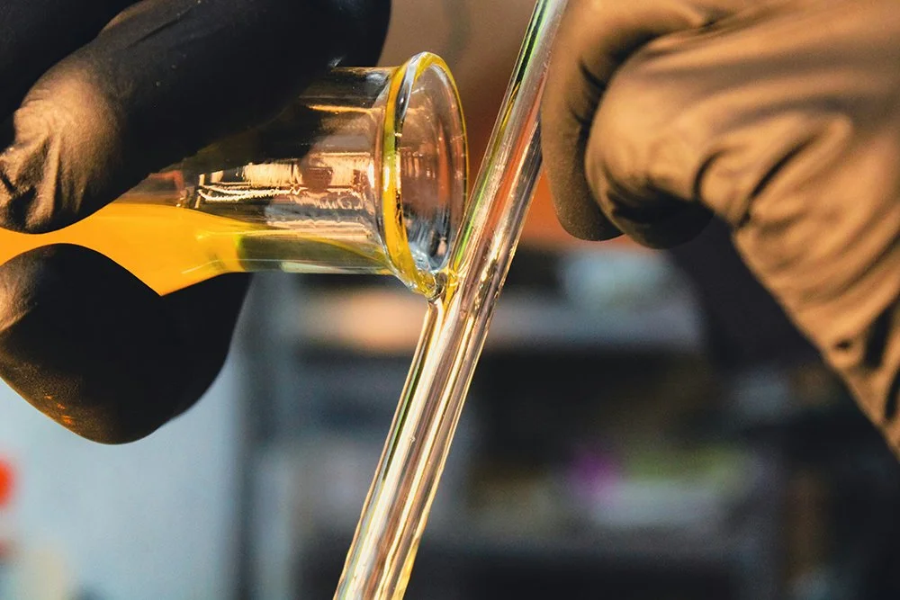 Persona con guantes de laboratorio vertiendo un líquido amarillo en un tubo de ensayo de vidrio.