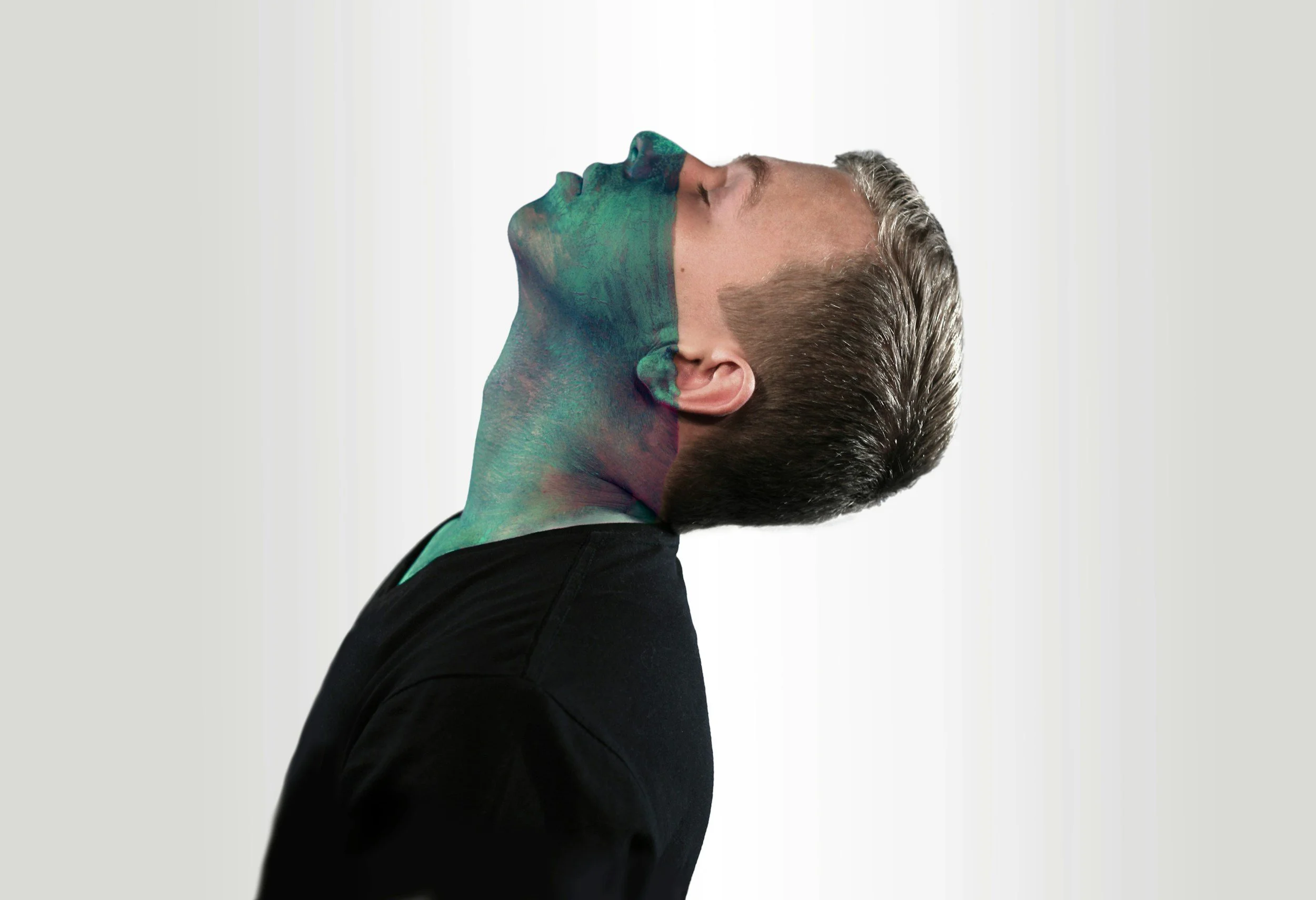 A man with his head tilted back, with a paint design on the left side of his face and neck, and wearing a black shirt as he looks upwards against a plain gray background.