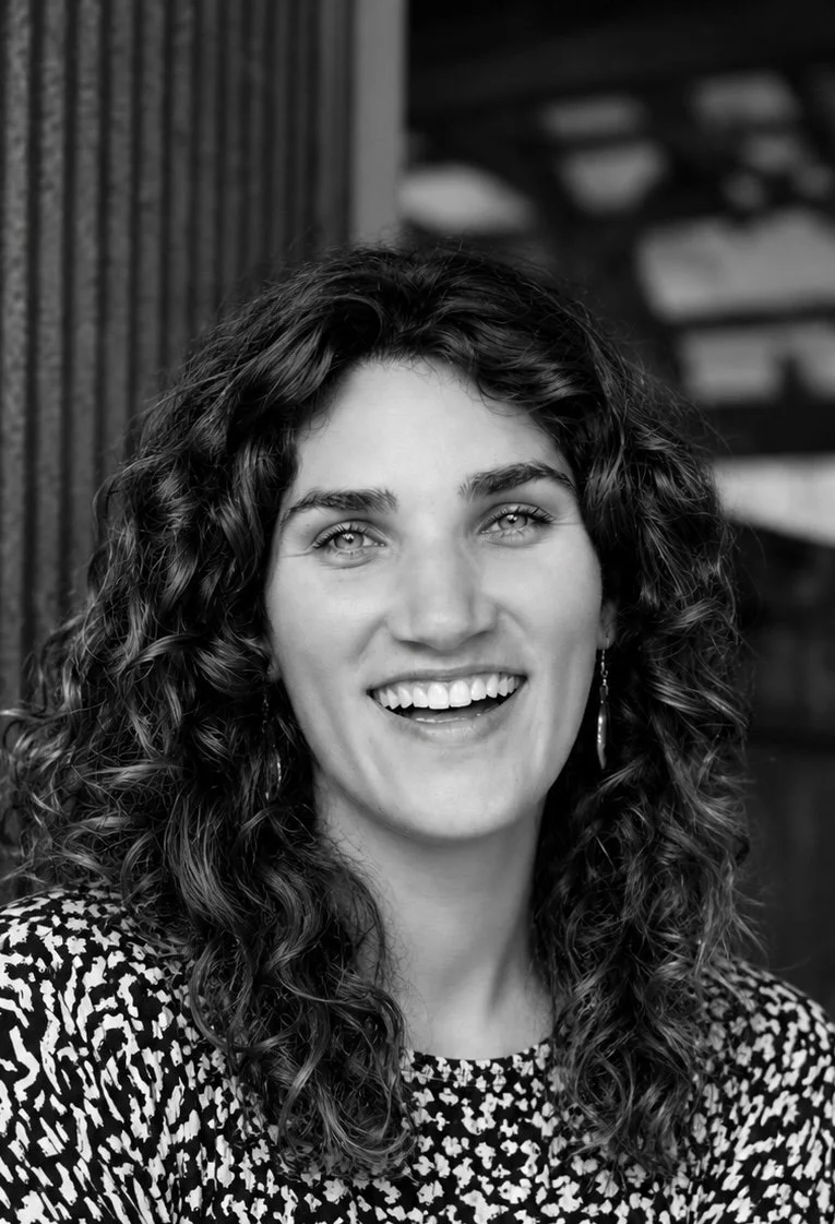 Black and white portrait of a young woman with curly hair, smiling, wearing earrings and a patterned top, in front of a textured wall.