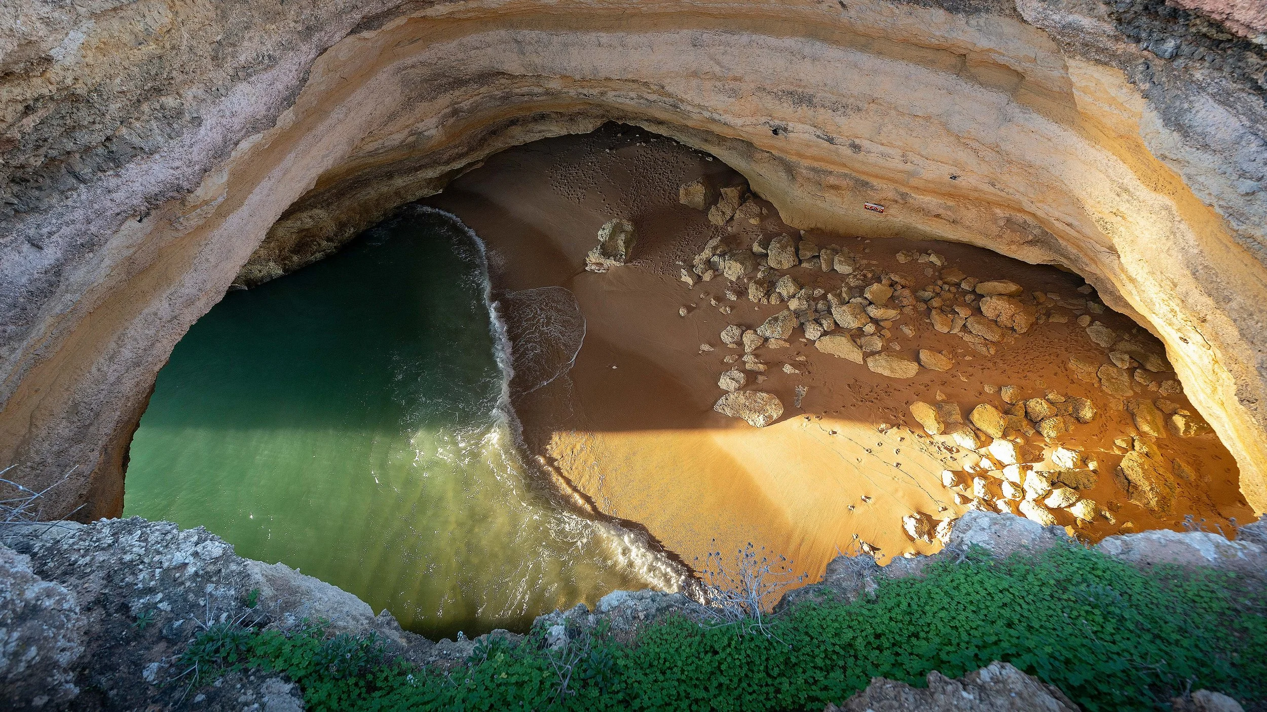 Aerial view of a coastal cave with a sandy beach and green water, surrounded by rocky cliffs.