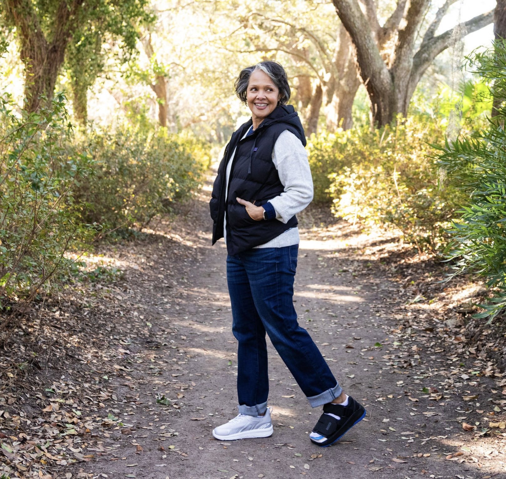 Woman walking on a dirt trail in a wooded park, smiling, wearing casual clothes and sneakers, with sunlight filtering through trees.