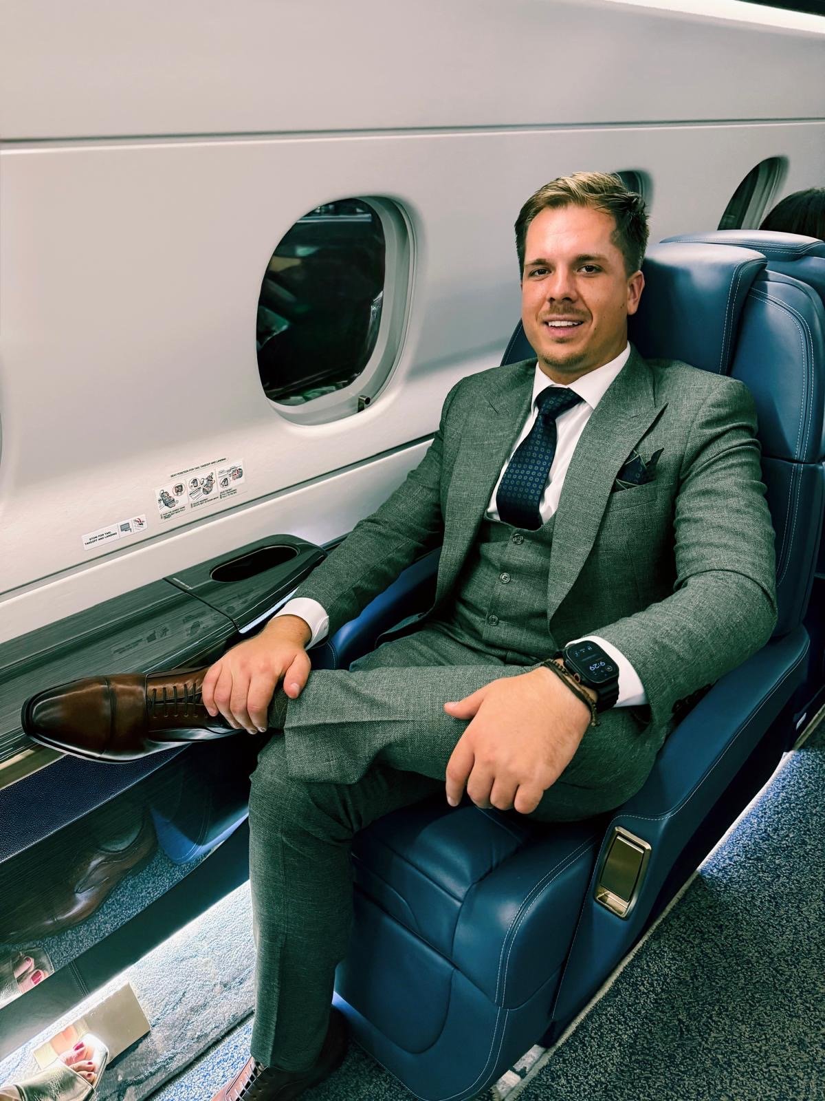 A man in a gray suit sitting in a premium airplane seat with a window beside him, smiling at the camera, wearing a smartwatch and holding his shoe.