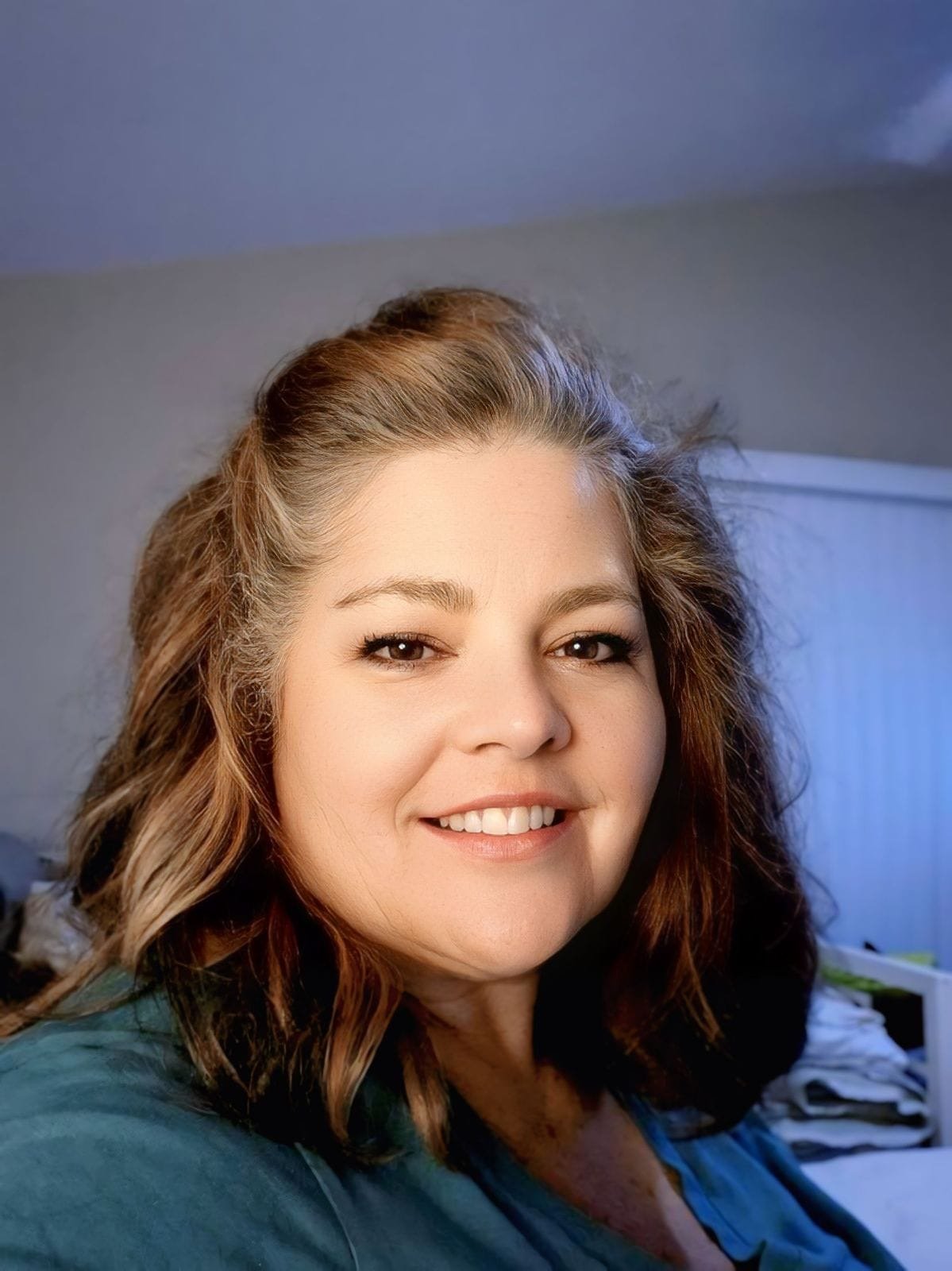 A woman with shoulder-length wavy brown hair and a smile, wearing a teal top, is indoors in a room with a blue and gray background.
