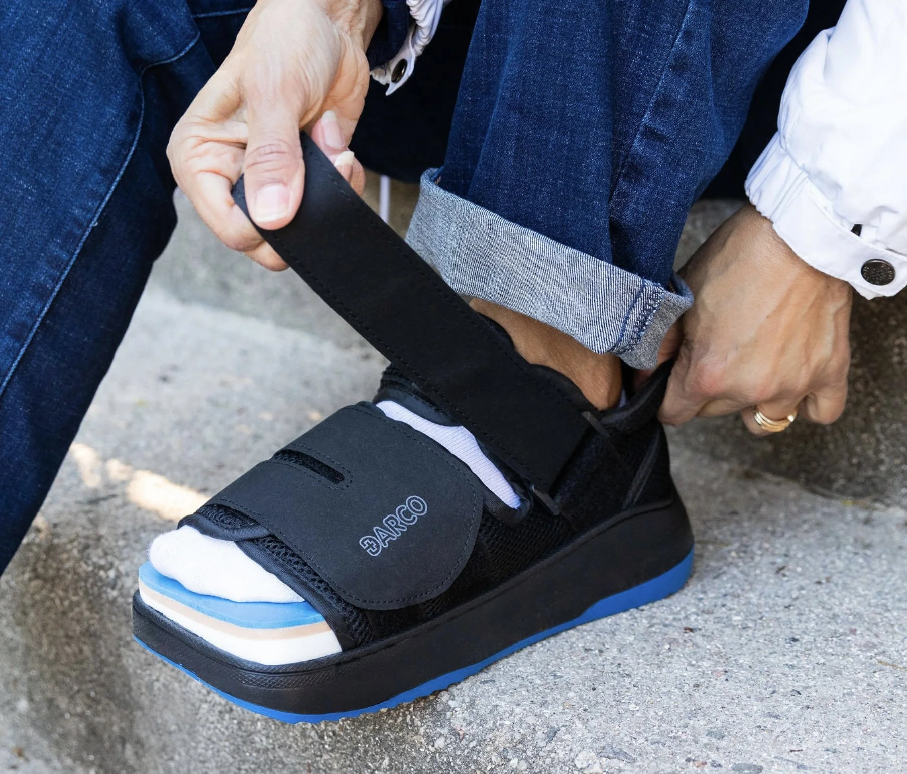 Person fastening a black ankle brace on their foot while sitting on a concrete surface, wearing jeans and a white jacket.