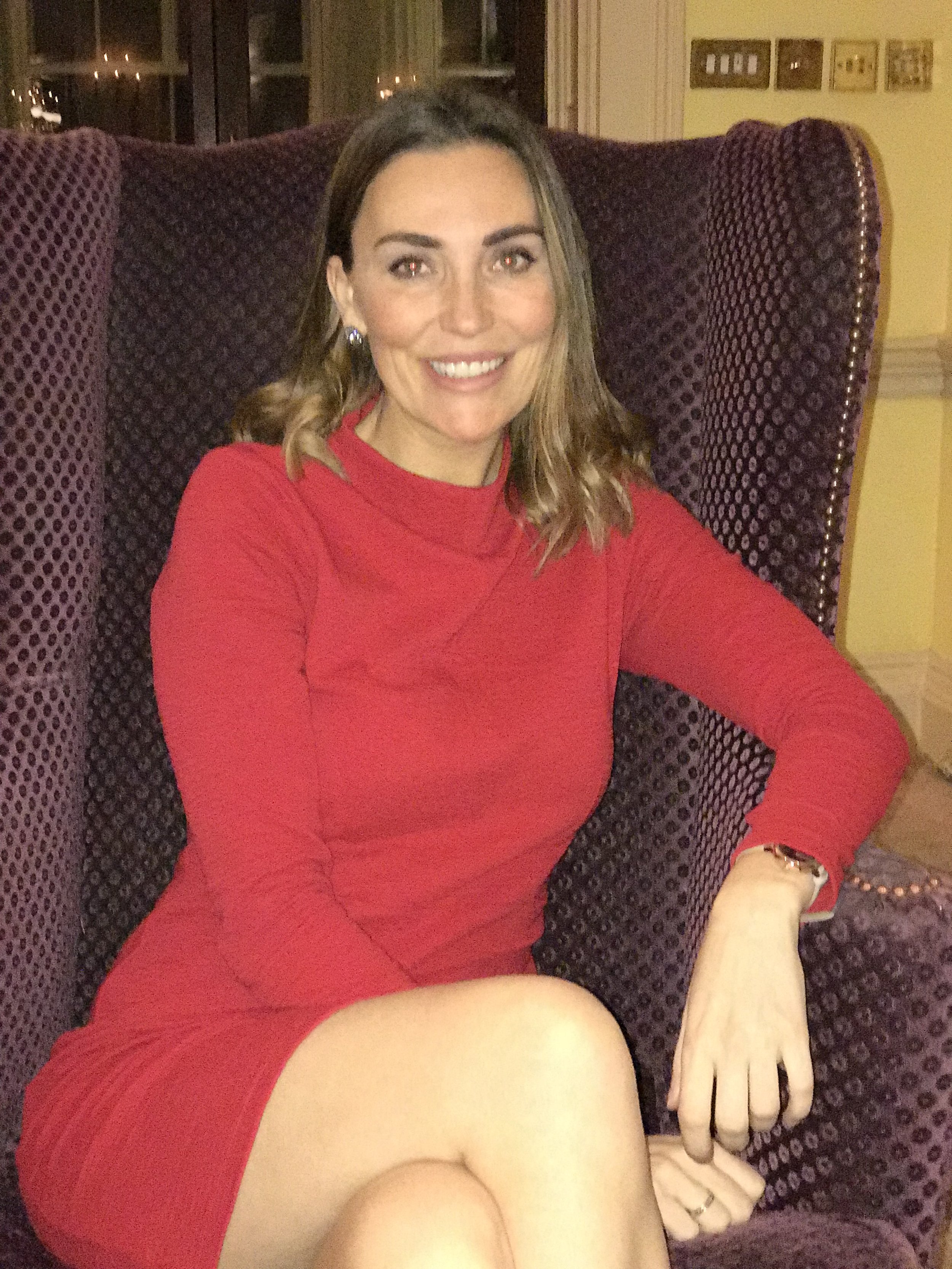 A woman with brown hair sitting on a purple, high-backed armchair inside a house. She is wearing a red dress and smiling at the camera.