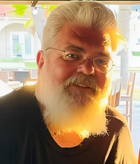 Man with gray hair, beard, and glasses, smiling in a sunlit outdoor setting