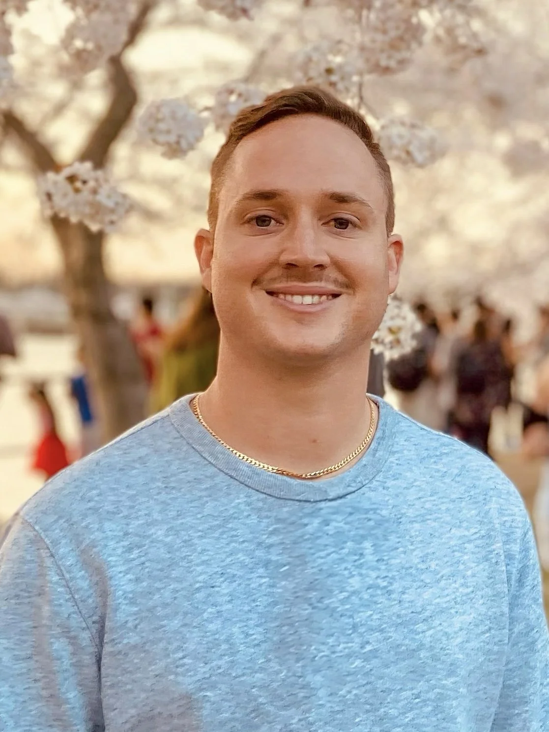 Smiling young man with short hair standing outdoors near blooming cherry blossom trees during sunset or golden hour.
