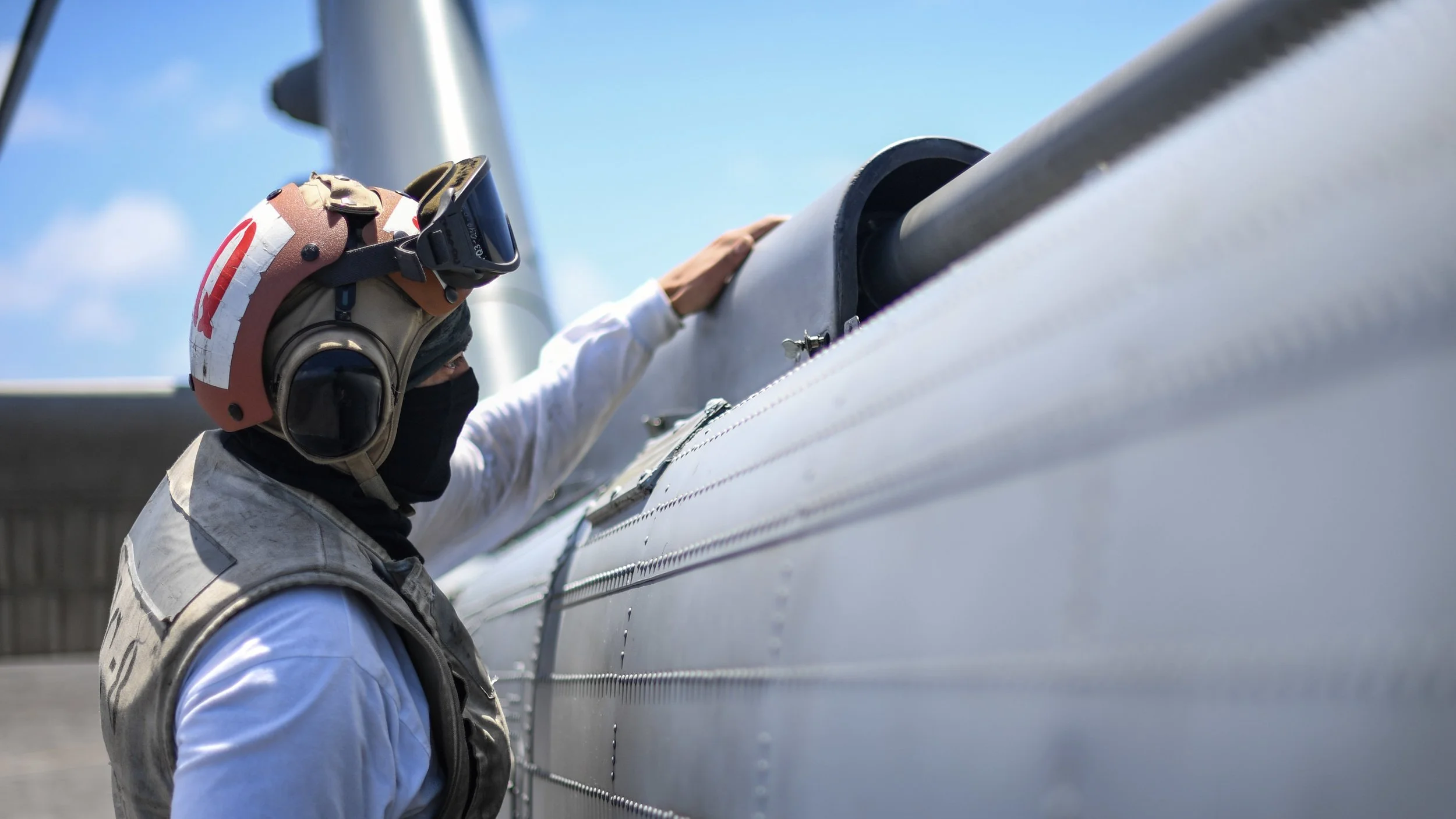 A person wearing a pilot's helmet, goggles, and a vest, inspecting or working on an aircraft fuselage outdoors under a blue sky.