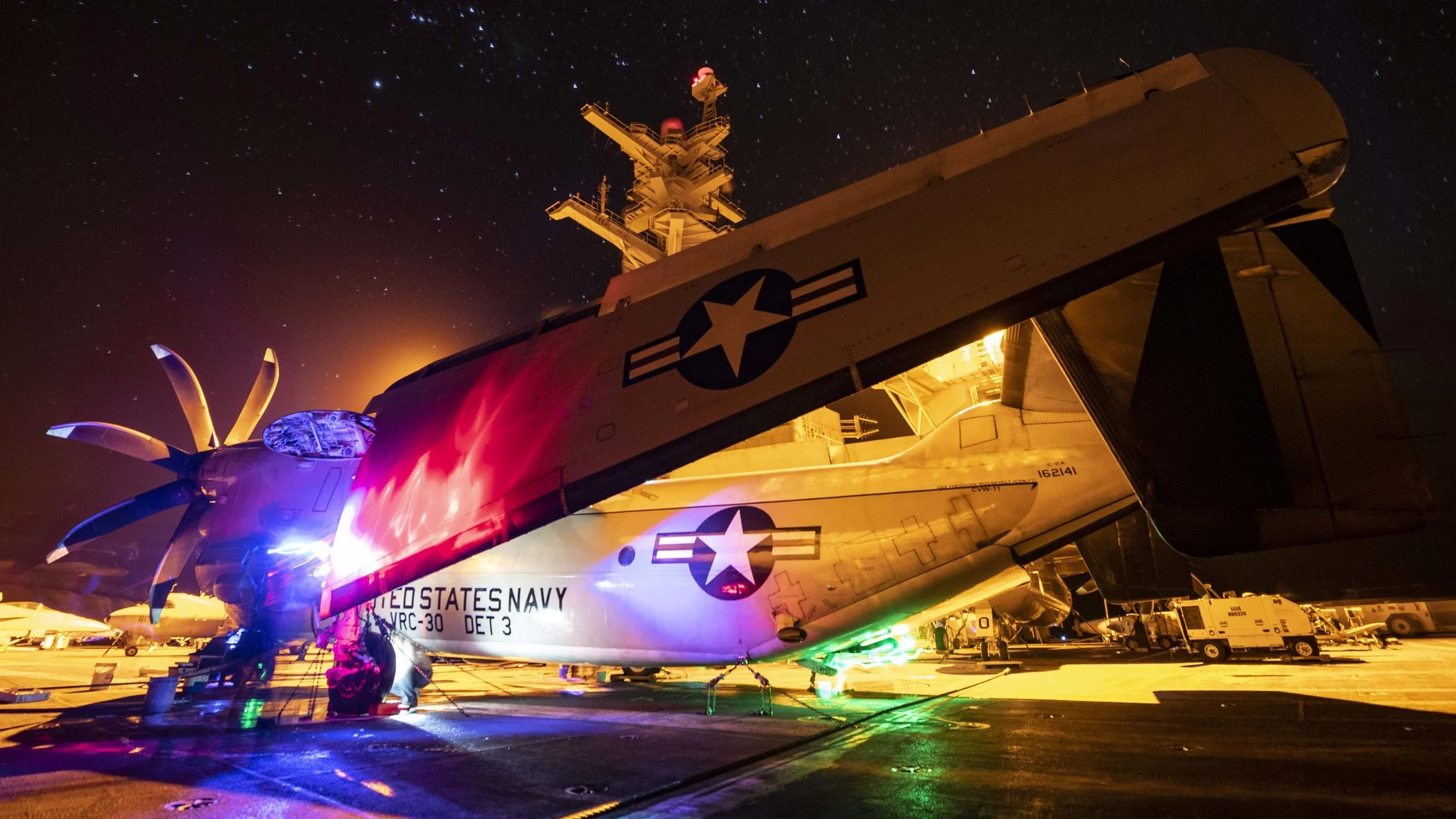 A U.S. Navy aircraft on an aircraft carrier at night, illuminated by colorful lighting and under a starry sky.