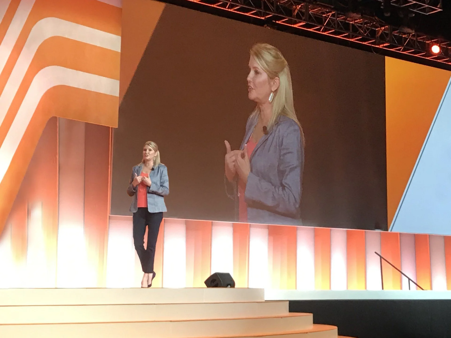 Pam Nemec is standing on stage with a large screen behind her showing a close-up of her speaking. The stage features orange and yellow decor, with a staircase and lighting setup.