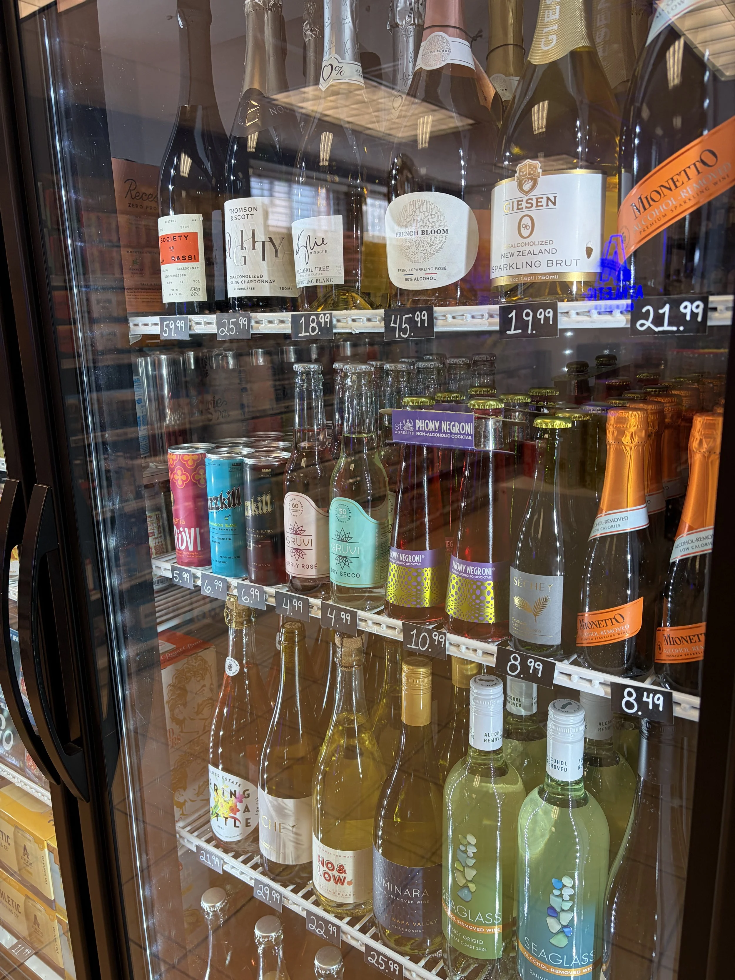 A refrigerated display case filled with various bottles of non-alcoholic wine and non-alcoholic beverages, with price tags visible on each shelf.