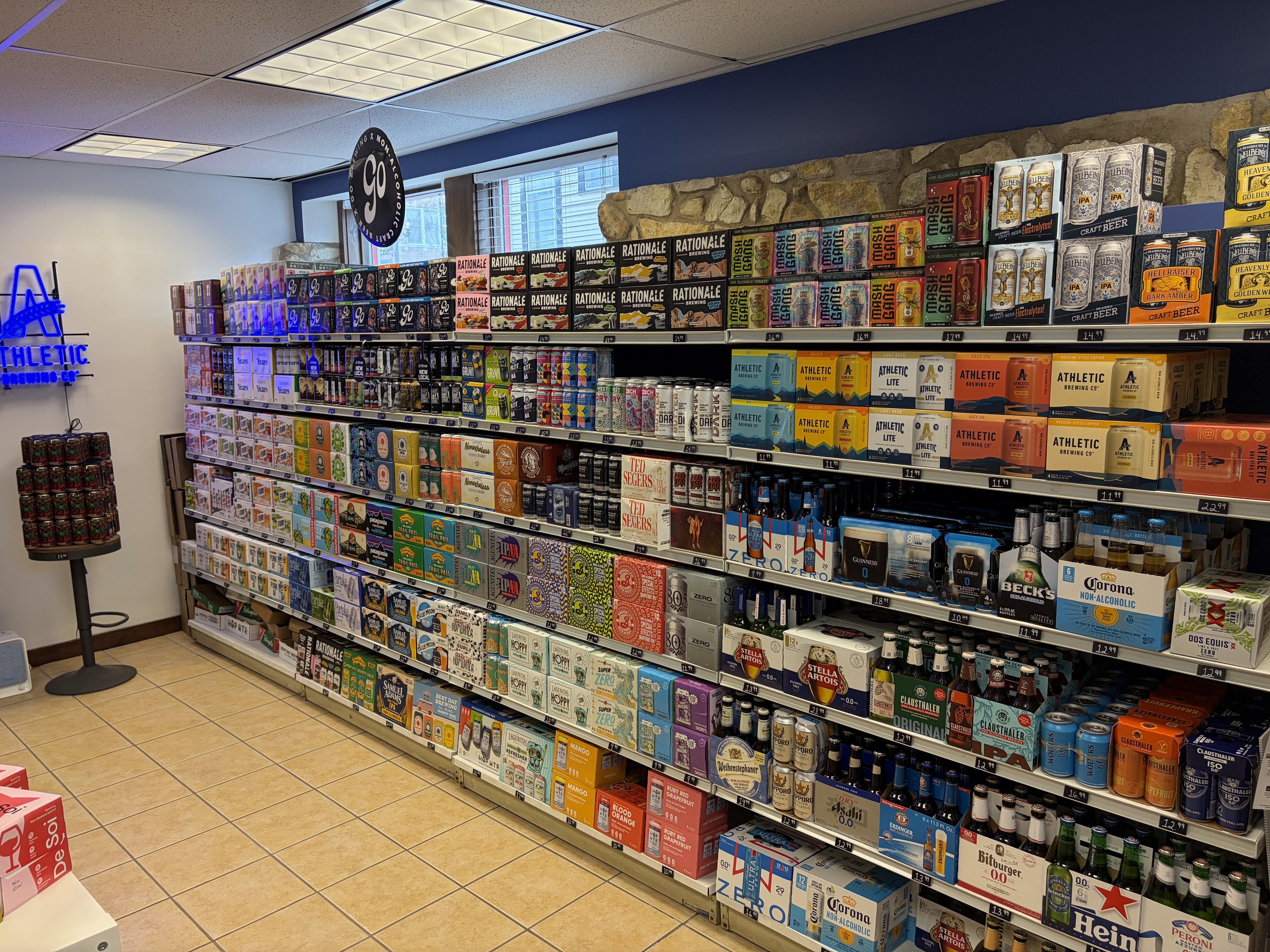 Store shelf stocked with various brands NA beer, including craft, domestic, and imported options, with visible prices. To the left, there is a display stand with cans of a different beverage, and a neon sign on the wall reads 'ATHLETIC BREWING CO.'