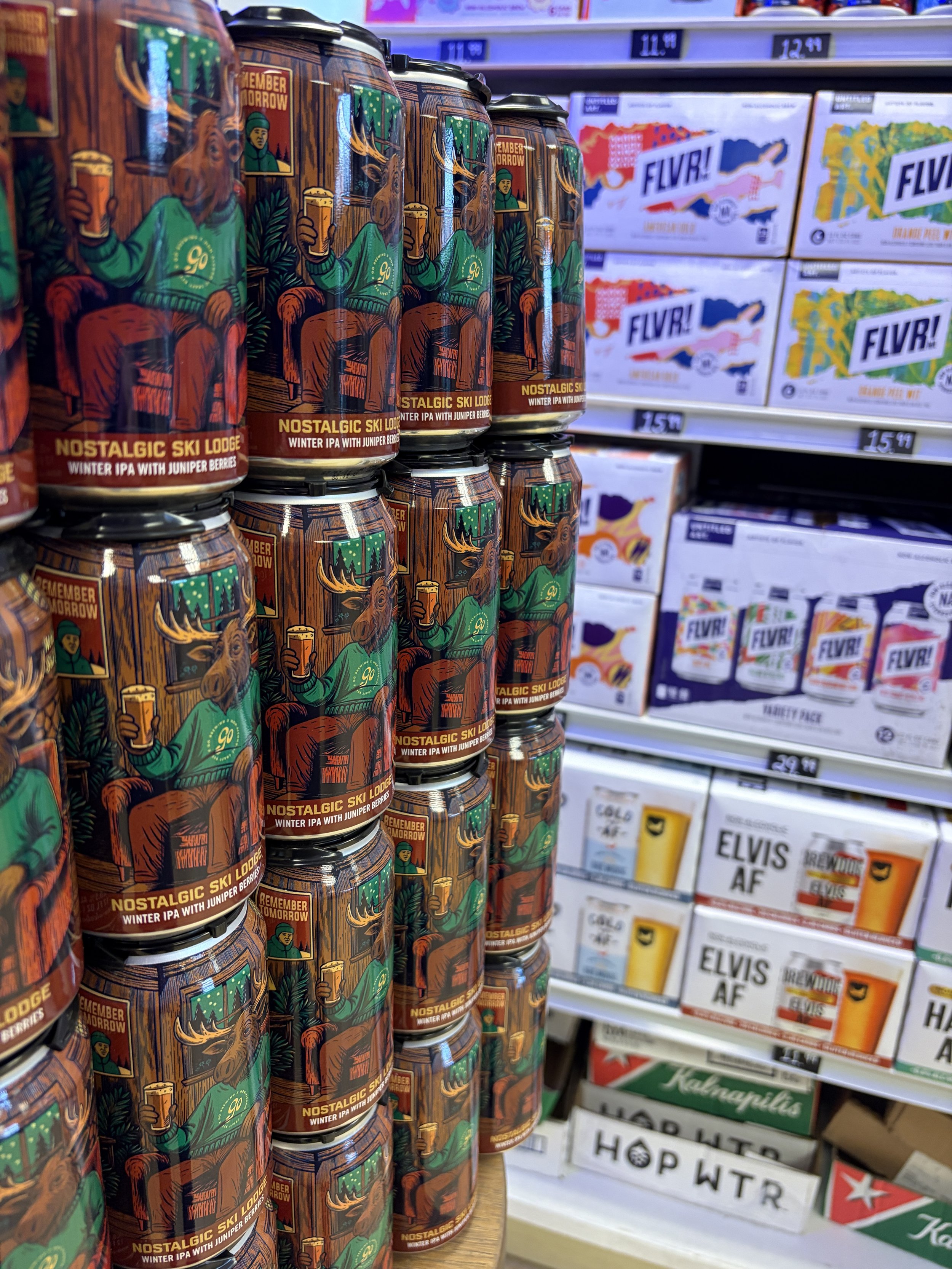 Cans of Nostalgic Ski Lodge Winter IPA with Juniper Berries NA Beer option stacked on a wooden shelf in a store. In the background, shelves with boxes of various non-alcoholic beverages including FLVR! and Elvis AF.