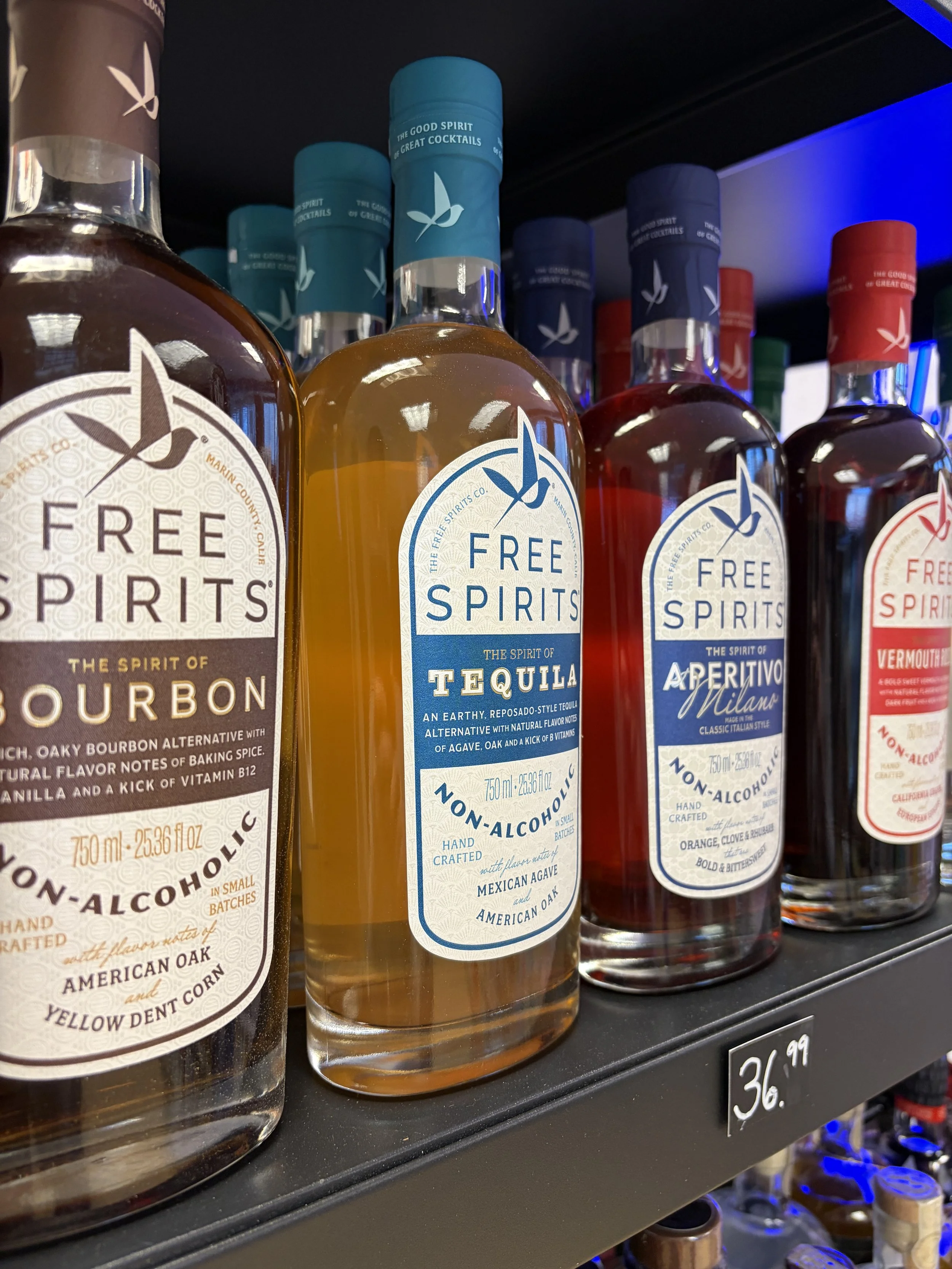 Bottles of non-alcoholic spirits on a store shelf, including brands like Free Spirits in flavors such as bourbon, tequila, and aperitivo, with a price tag of $36.99.