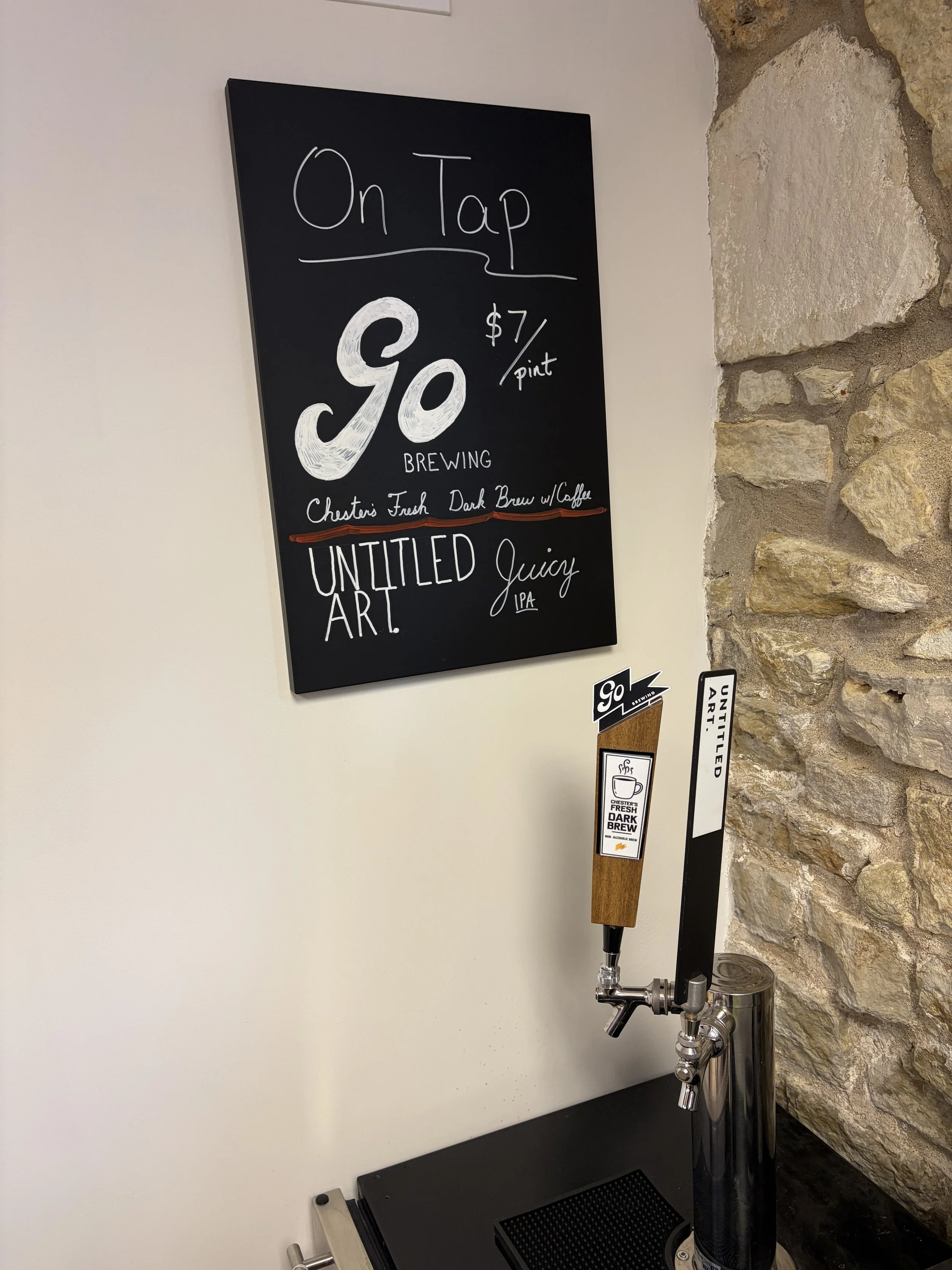 A black chalkboard sign advertising taproom offerings, with white and red writing. It mentions $7 per pint, features 'Go Brewing,' and lists 'Chester’s Fresh Dark Brew with Coffee' and 'Untitled Juicy IPA.' There is a beer tap dispenser below the sign with labels reading 'Go' and 'Untitled Art,' and a notice for Chester's Fresh Dark Brew.