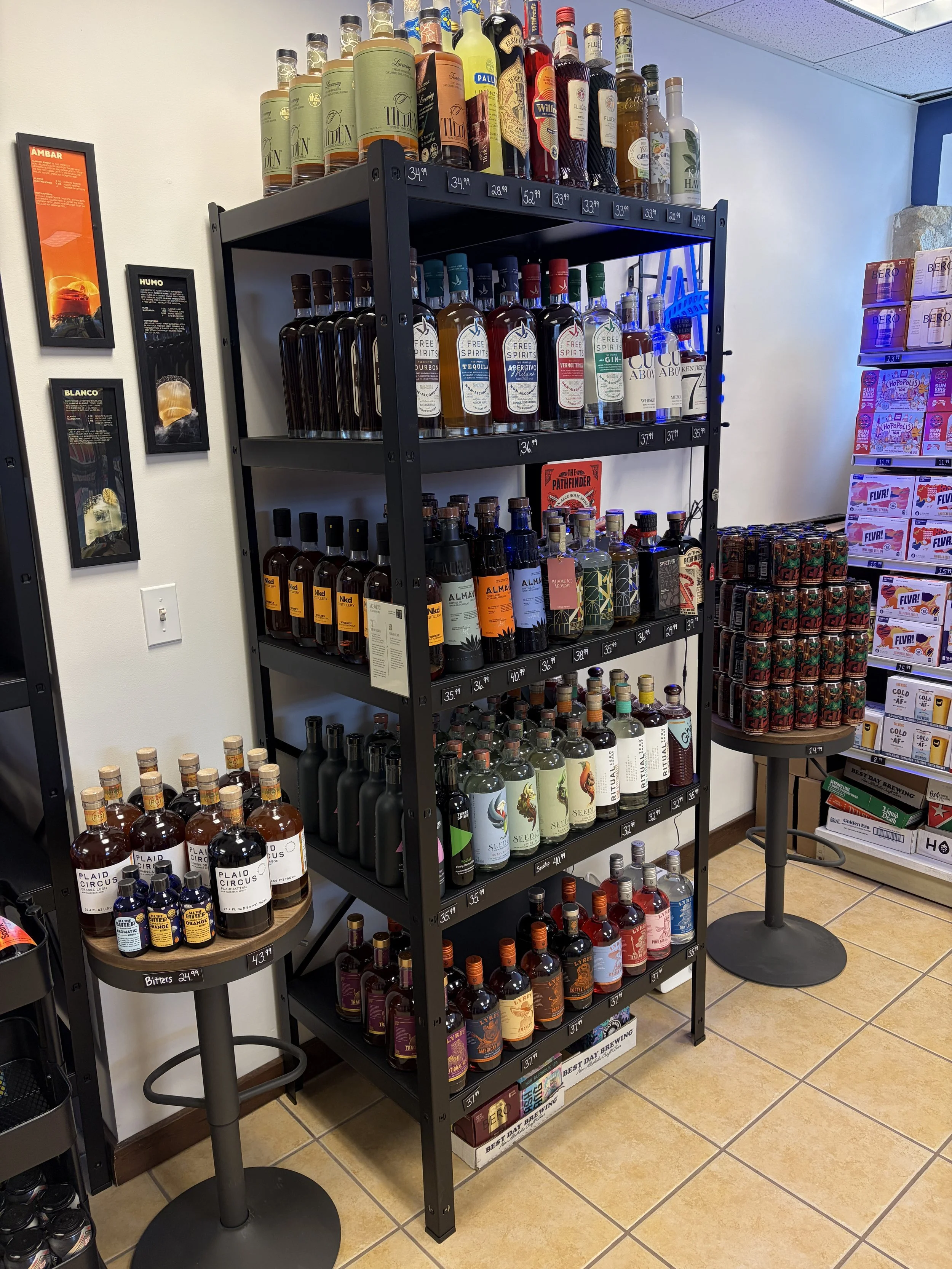 Display of various bottles of non-alcoholic spirits on black metal shelves in a liquor store, with some bottles on small round tables nearby.