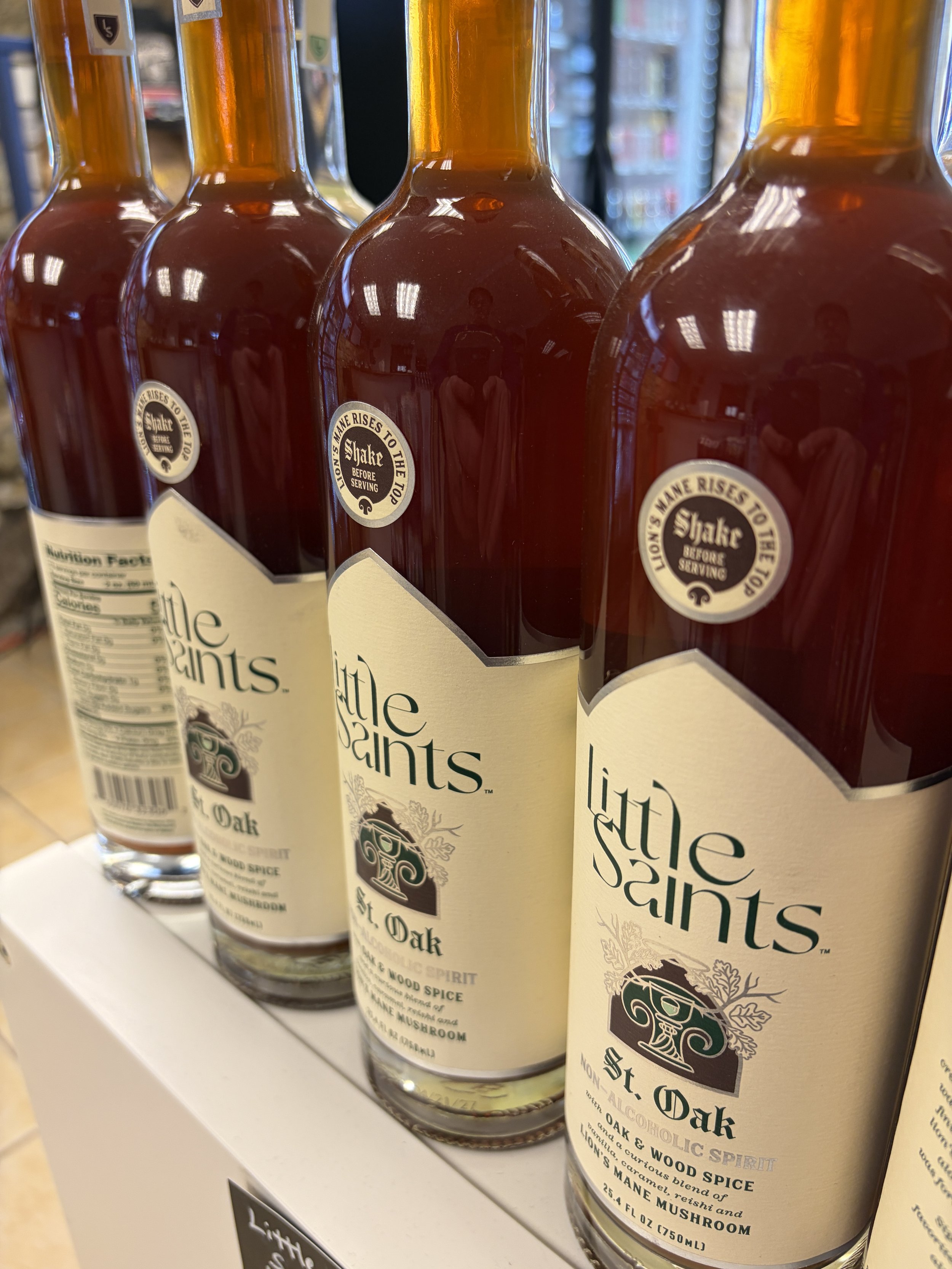Shelves with bottles of Little Saints St. Oak non-alcoholic spirit, with labels indicating ginger, oak, and wood spice flavor, and instructions to shake before serving.