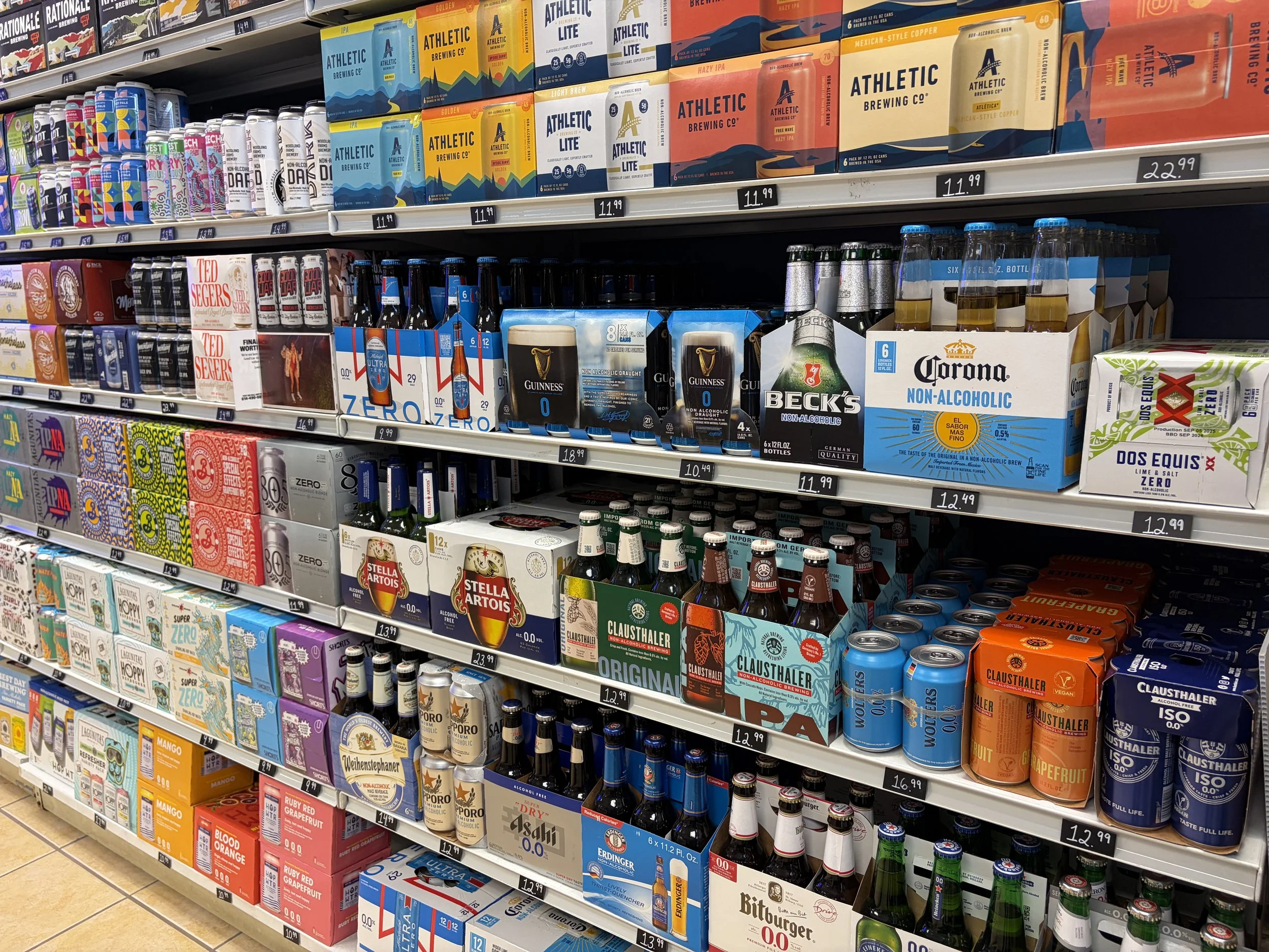 Downtown wheaton bottle shop shelf with non-alcoholic beverages, including selections from brands like Athletic Brewing, Guinness, Beck's, Corona, Dos Equis, Stella Artois, Clausthaler, and others. The prices are displayed on tags below each product.