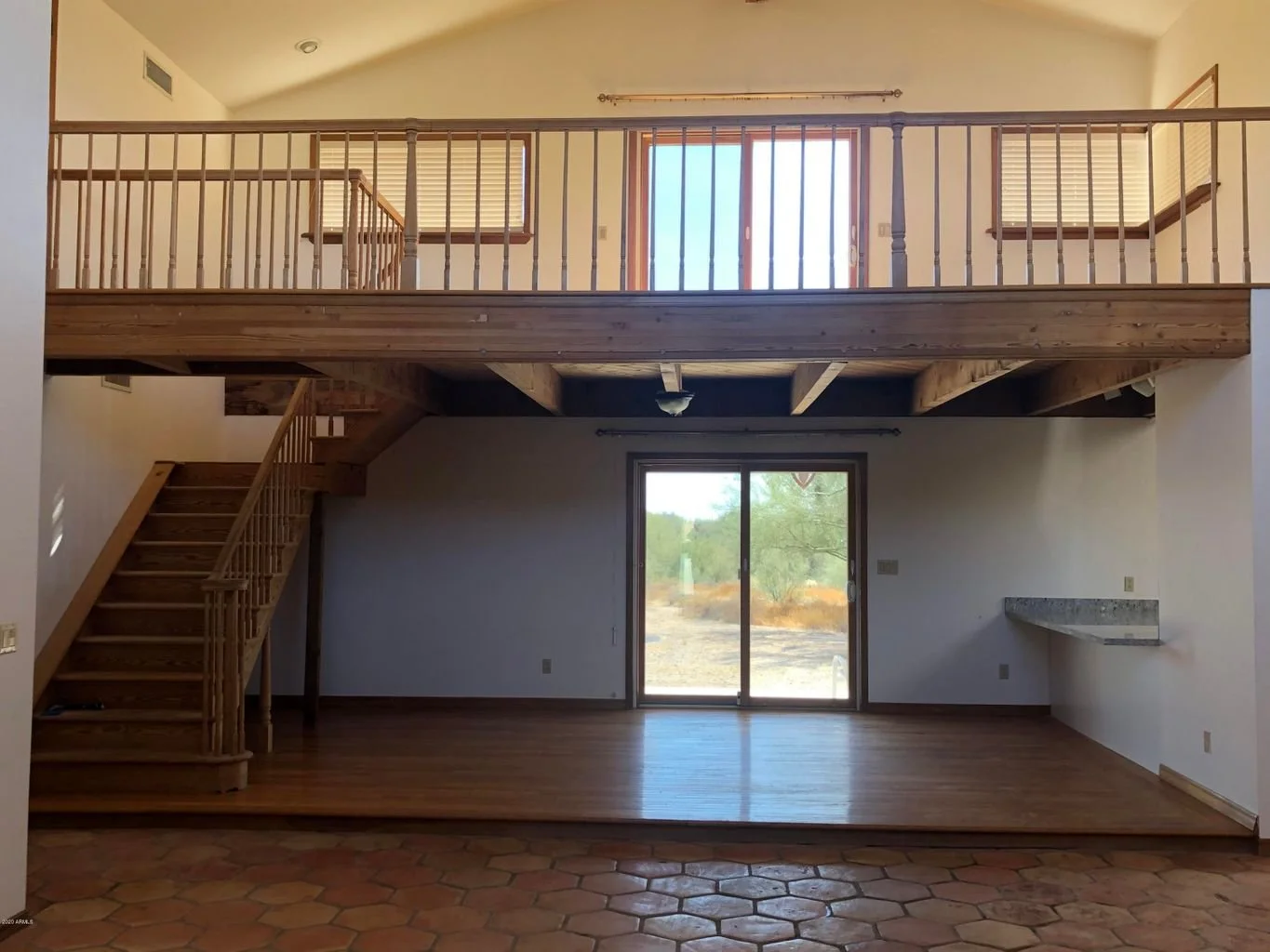 Interior view of a two-story house with a wooden staircase leading to a second-floor balcony overlooking the living area. Large glass sliding doors open to an outdoor landscape, and a small built-in countertop is on the right wall. The ceiling is hig