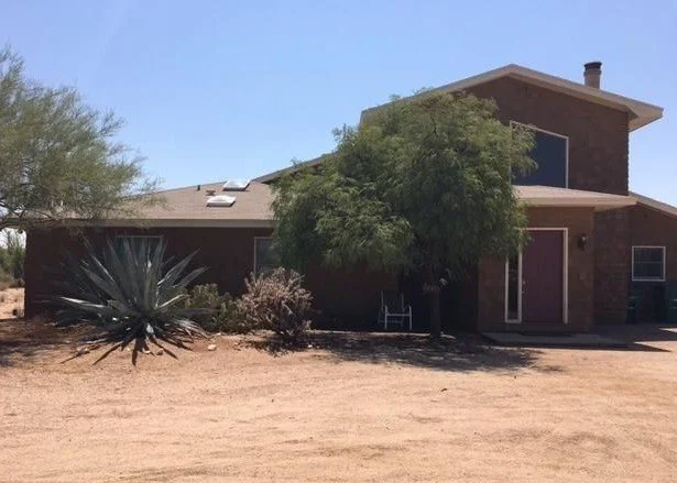 A two-story brick house with large windows and a front porch, surrounded by desert landscaping and a few trees.