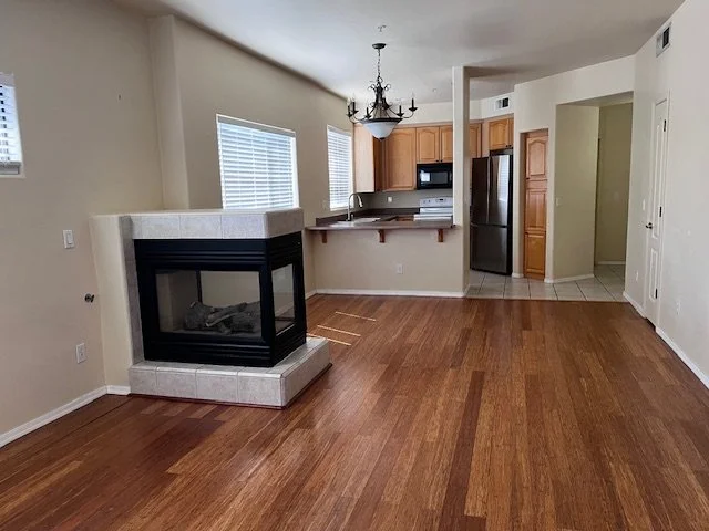 Empty living room with hardwood floors, fireplace, kitchen with wooden cabinets, stainless steel appliances, and a chandelier.