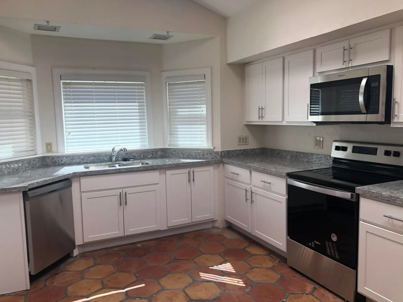 Kitchen with white cabinets, gray speckled countertops, stainless steel appliances including microwave, oven, and dishwasher, three windows with closed blinds, and terracotta tile flooring.