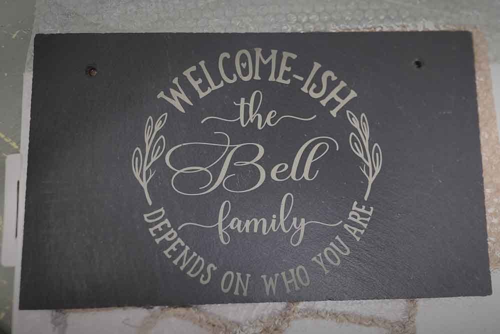 A decorative black plaque with white cursive and block lettering that reads, "Welcome-ISH the Bell family. Depends on who you are," on a stone surface background.