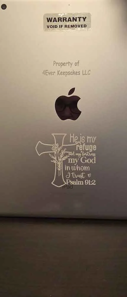 The back of a computer or electronic device with a warranty sticker saying "VOID IF REMOVED," a logo of Apple, and a decorative cross with a Bible verse from Psalm 91:2 that says, "He is my refuge and my fortress, my God in whom I trust."