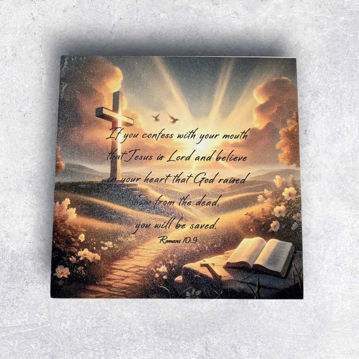 Religious artwork with a cross on a hill, open Bible, and sunset sky with clouds, birds, and rays of light, featuring a quote from Romans 10:9 about confessing Jesus and being saved.