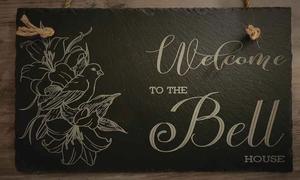 Black welcome sign with white floral outline and bird illustration reading "Welcome to the Bell House" mounted on a wooden surface.