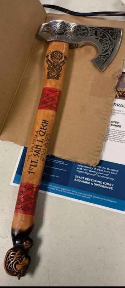A decorative axe with a wooden handle, engraved details, red wrapping near the top, and black designs at the bottom. It is resting on a cardboard box next to some papers and a pamphlet.