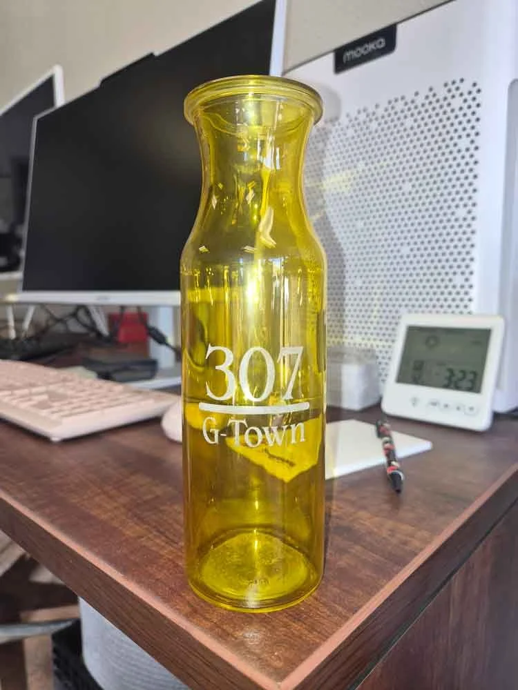 Yellow glass bottle with white text reading "30% G-Town" placed on a wooden desk with computers, a clock, and a pen in the background.