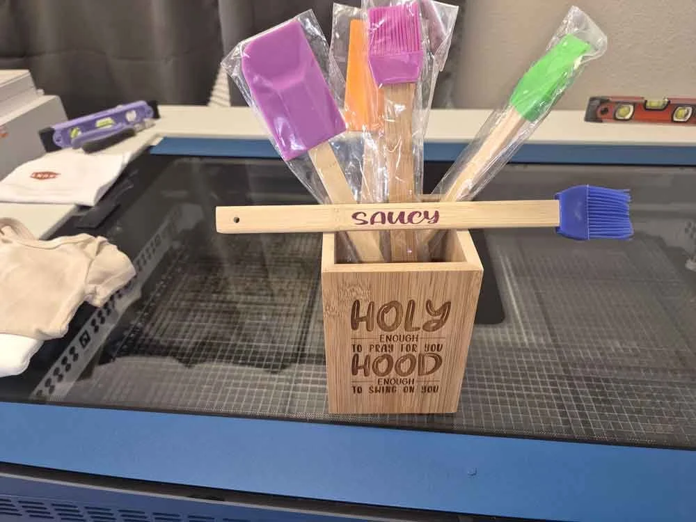 A wooden container with the words "Holy Hood" engraved on it, holding colorful silicone basting brushes with wooden handles and plastic bristles.