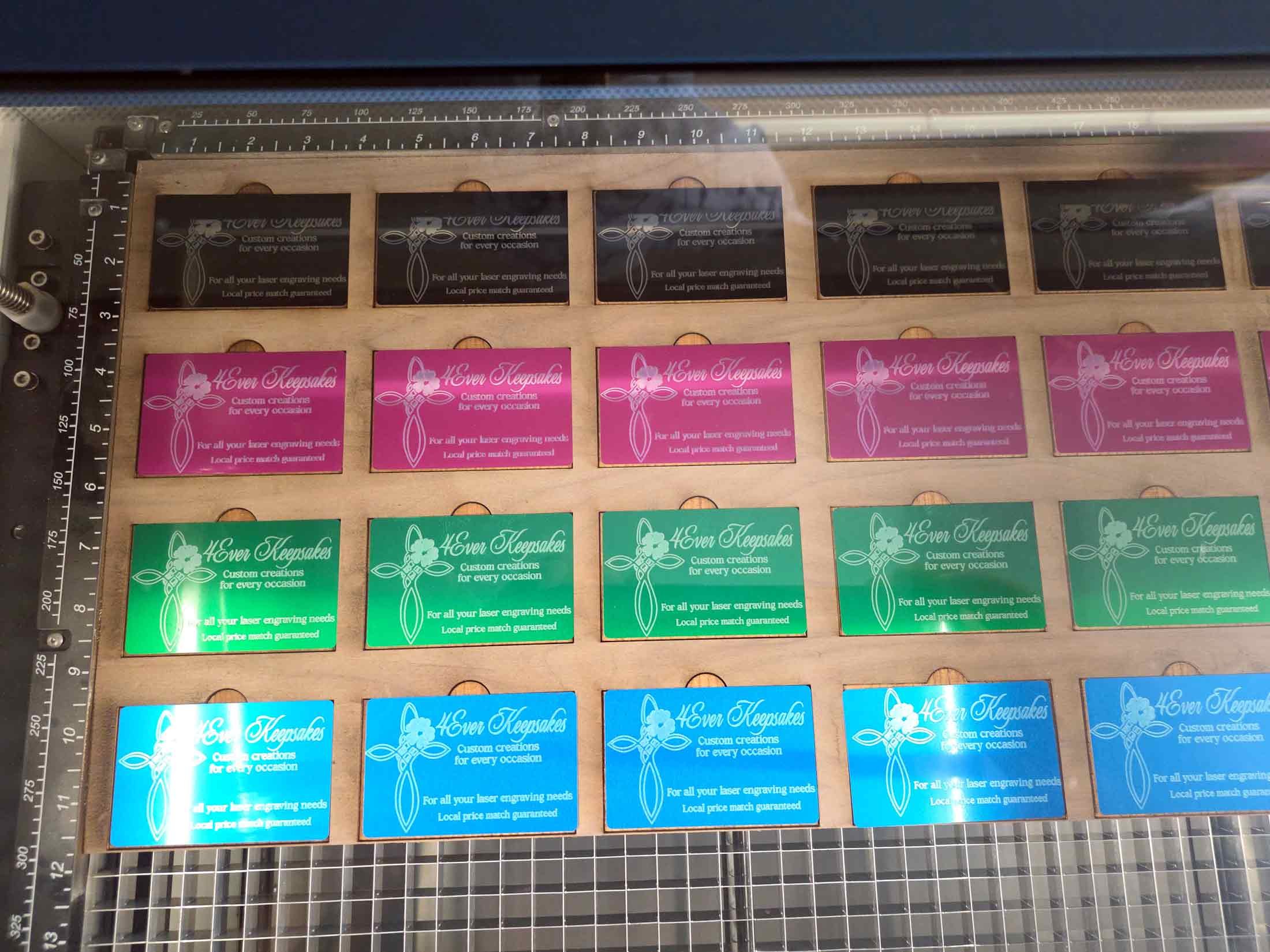 Colorful laser-engraved acrylic business cards in black, pink, green, and blue, arranged in rows on a wooden board inside a laser cutter, with measurement grid and metal clamps visible.