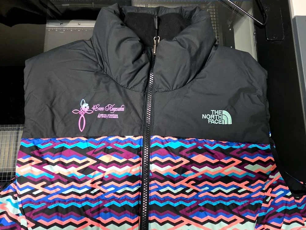 A black The North Face puffer jacket with a colorful geometric patterned section in pink, purple, blue, and black, with logos for 'Love Keepsakes' and 'The North Face'.