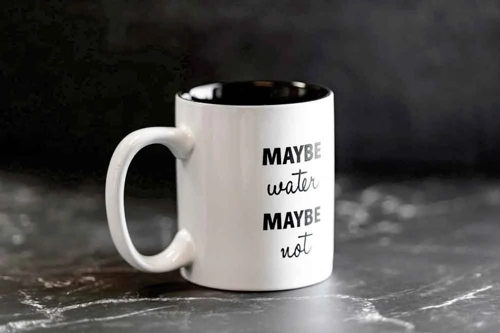 A white coffee mug with black text that reads "Maybe water maybe not," placed on a dark surface with a dark background.