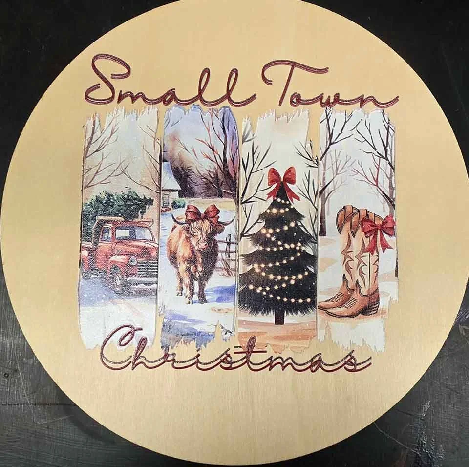 Round decorative holiday sign with the words 'Small Town Christmas' and four vertical panels featuring winter scenes, a Christmas tree with lights and a bow, a cow with a red bow, a vintage truck, and cowboy boots with a bow.