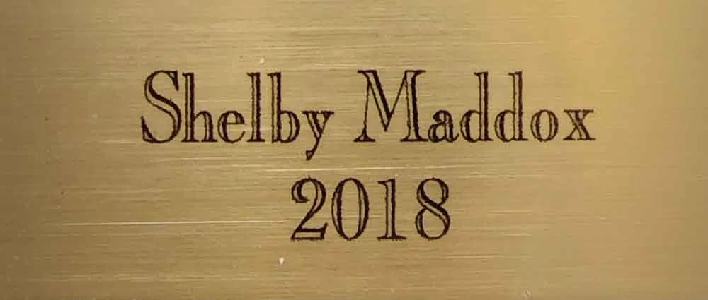 Close-up of a metallic plaque with engraved text reading 'Shelby Maddox 2018' on a golden background.