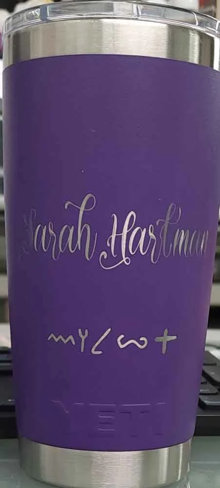 A purple travel mug with silver accents that has the name Sarah Hartman and decorative symbols, placed on a surface with a blurred background.
