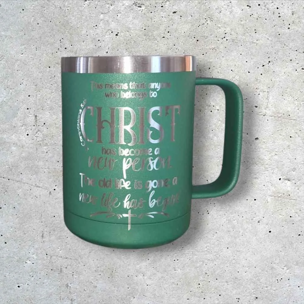 A green insulated mug with a silver rim and handle, featuring religious text and symbols, placed on a gray concrete wall background.