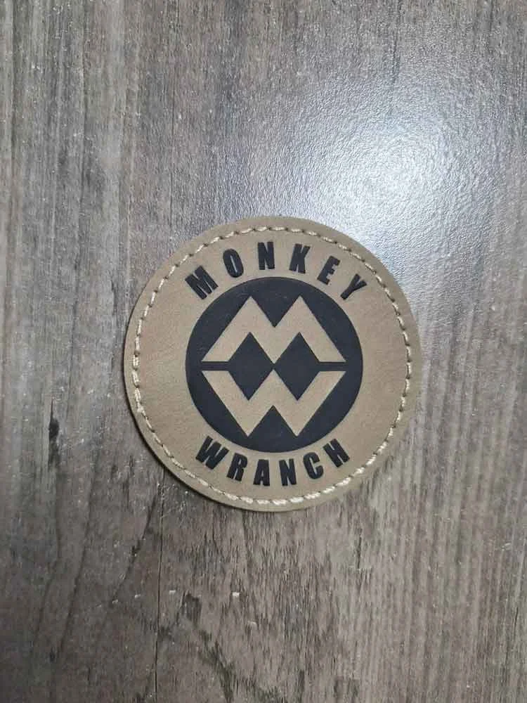 Leather patch with the words 'Monkey Wrangler' and a stylized logo of two overlapping monkeys on a wooden surface.