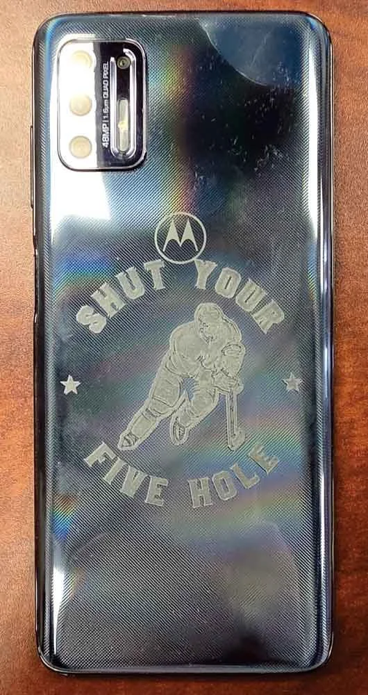 Back of a Motorola smartphone with a holographic finish, featuring a triple camera setup, a logo, and a reflective engraved sticker that says 'PUT YOUR FUE HOLLE' with an illustration of a person skating or snowboarding.