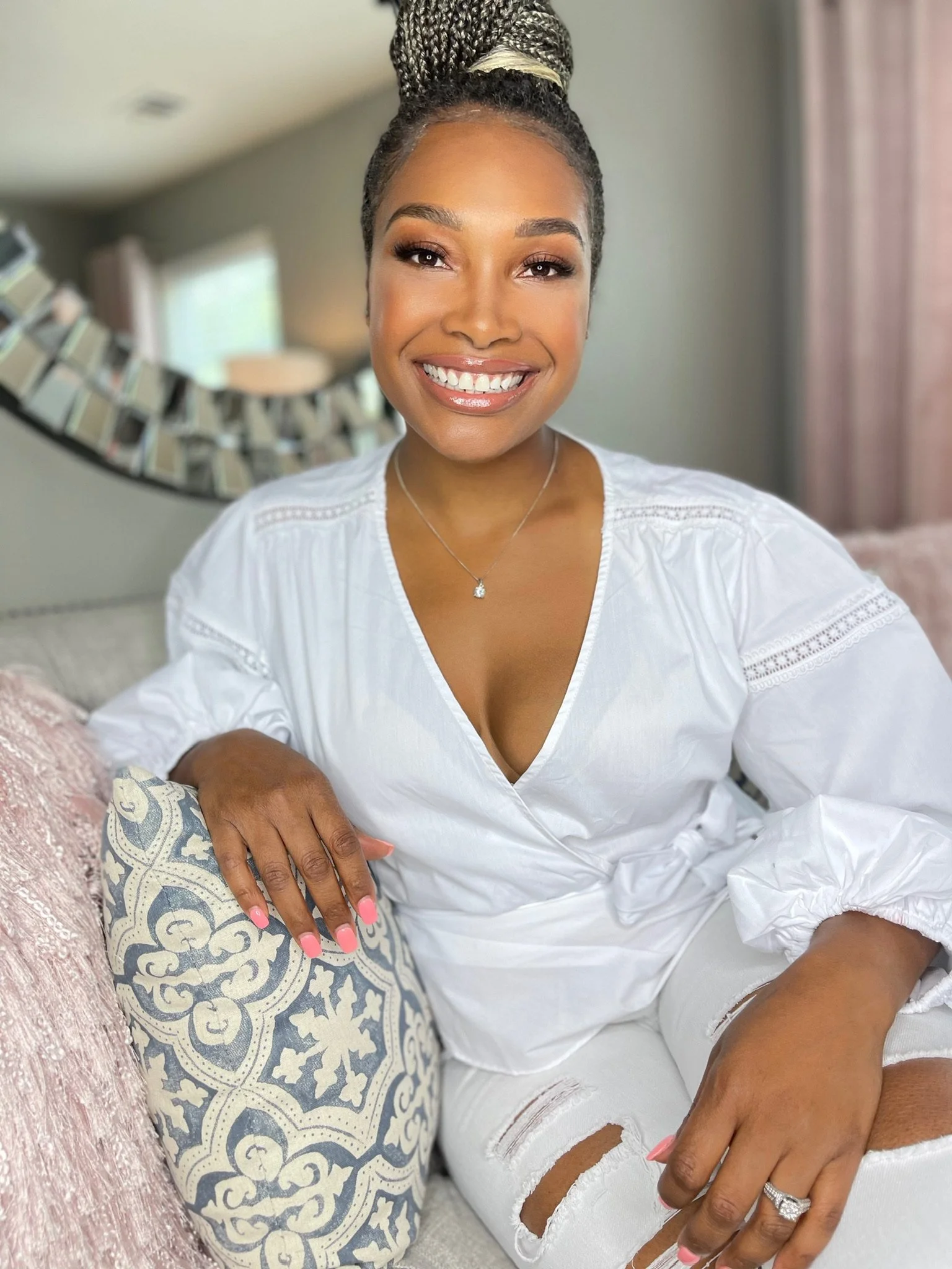 Akeila Goolsby, life coach and tech professional, in a warm lifestyle photo with braided hair styled in a top bun, wearing a white blouse and ripped jeans, sitting on a sofa, smiling at the camera representing the AkeilaB brand.