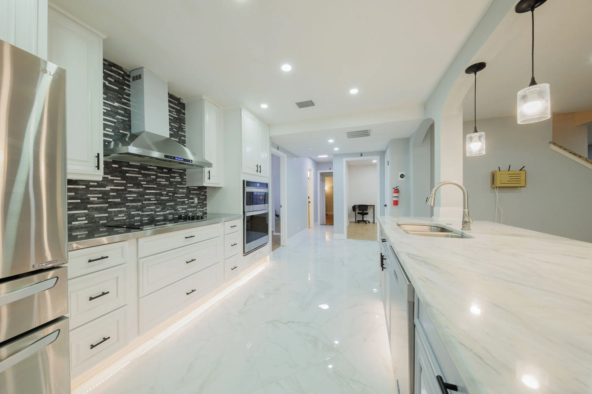 Modern kitchen with white cabinets, stainless steel appliances, black and gray mosaic backsplash, marble countertops, and pendant lights.