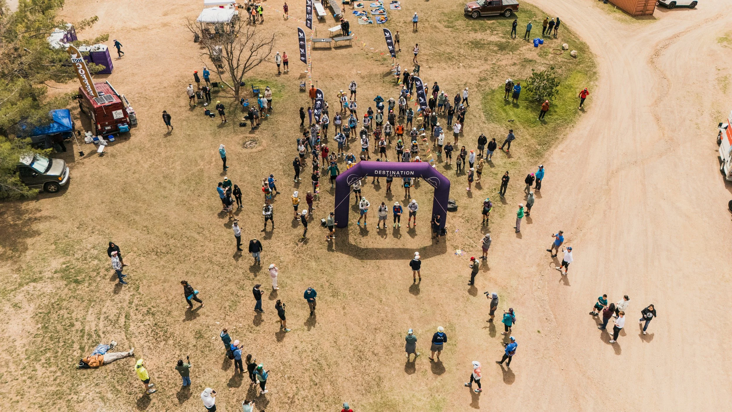 An aerial view of people gathering outdoors for a trail running event, with an inflatable arch at the start line labeled 'Destination Trail' and various tents and vehicles nearby.