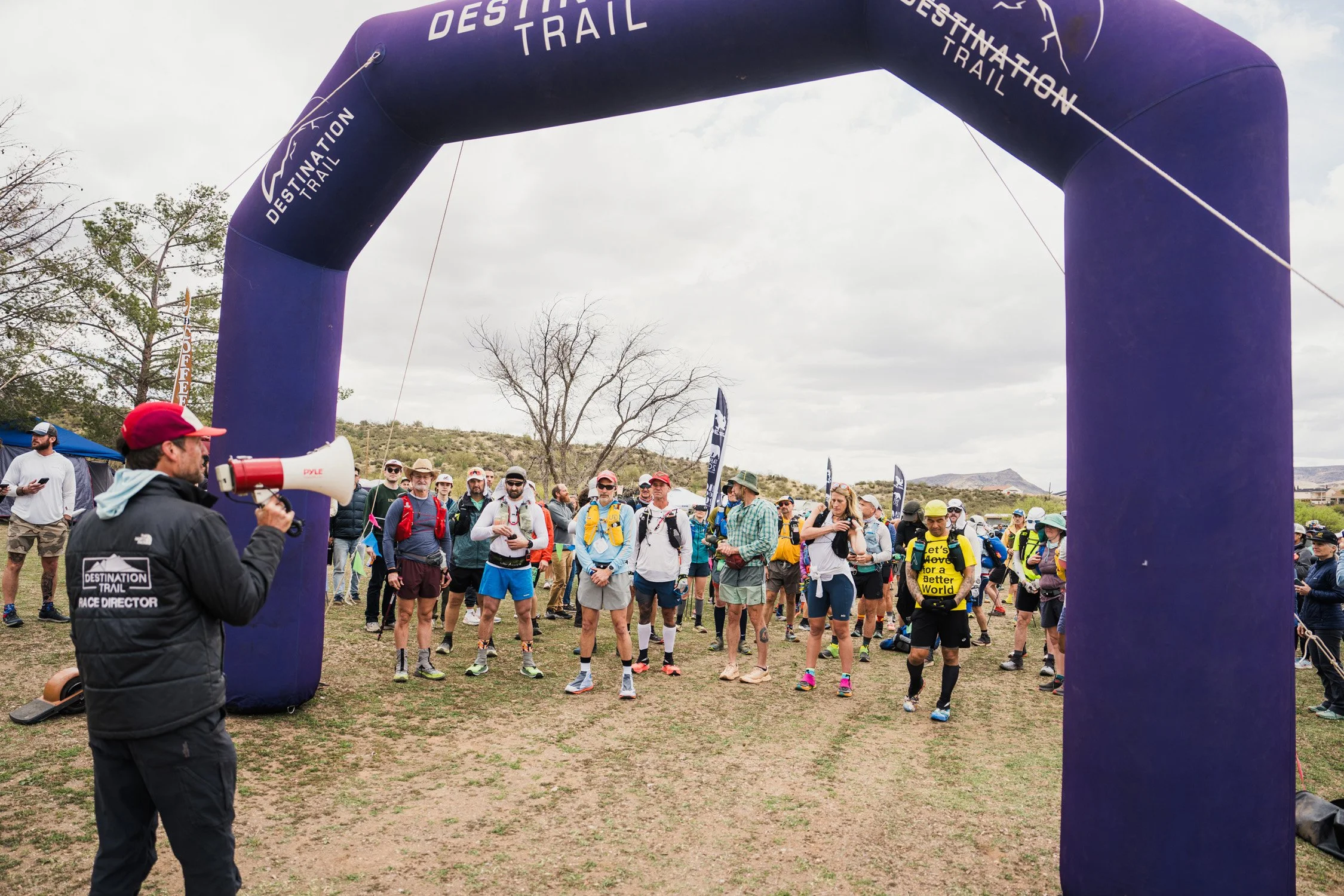 Group of people gathered at the start of a trail race under a large purple archway labeled "Destination Trail." One person in a black jacket with a megaphone is speaking to the crowd.