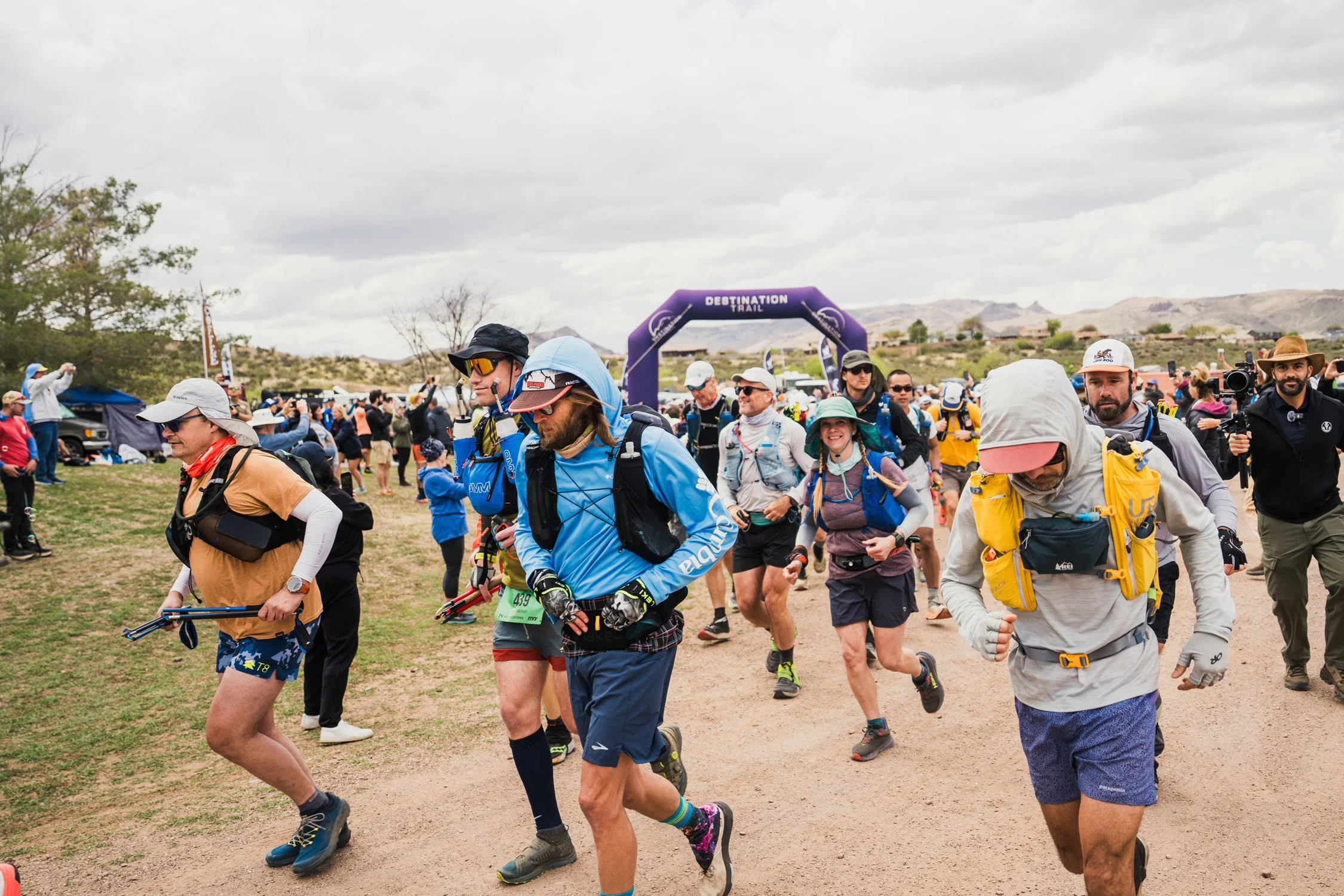 Group of people participating in an outdoor trail running event, starting or ending at an archway that reads 'Destination Trail,' with hills and cloudy sky in the background.