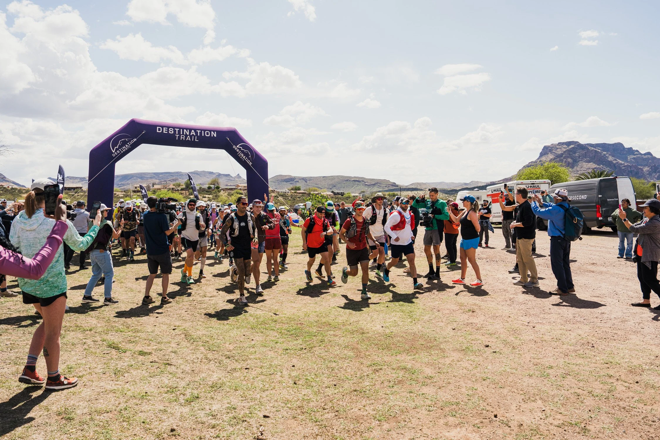 People participating in an outdoor race event, many wearing athletic clothing and backpacks, passing under a purple archway that reads 'Destination Trail' with a scenic landscape and mountains in the background.