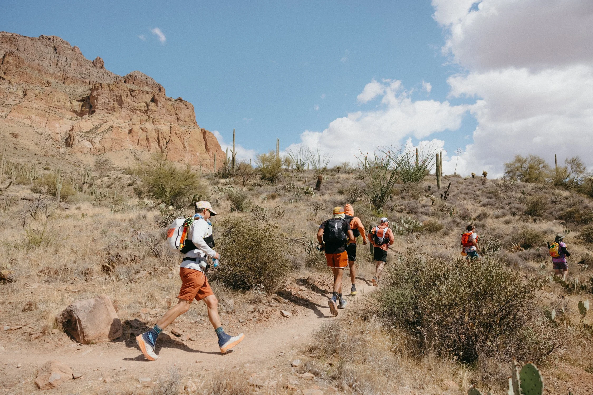 A group of hikers wearing backpacks and athletic clothing walking on a trail in a desert landscape with cacti, dry bushes, and rocky hills under a partly cloudy sky.