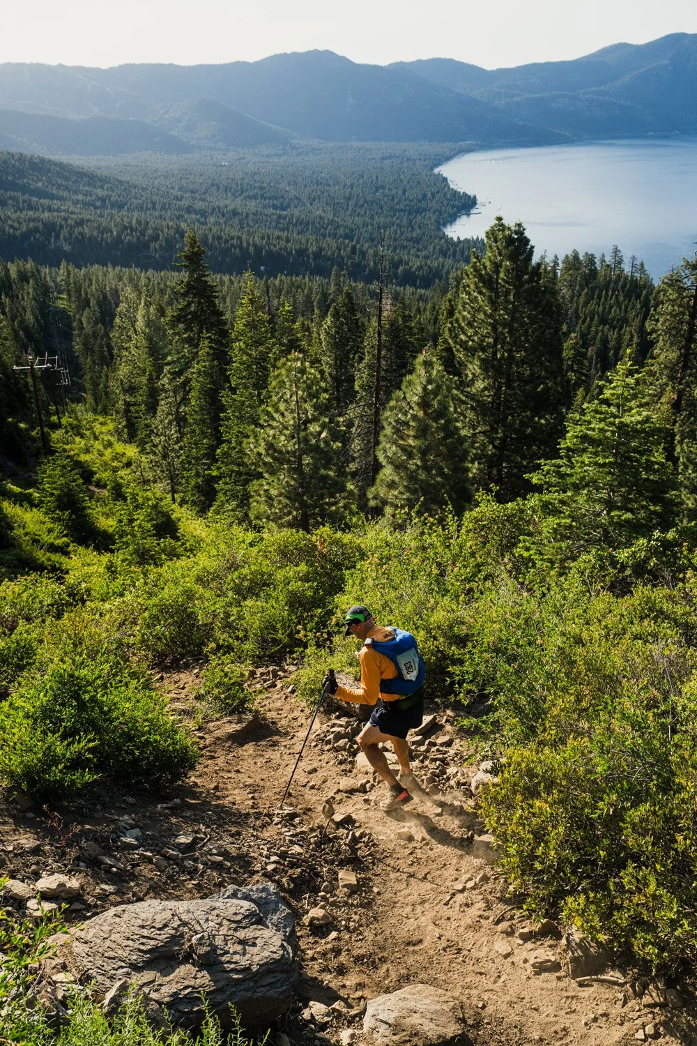 A person hiking downhill on a dirt trail through a green forest with a lake and mountains in the background.